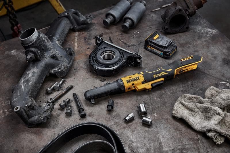 Herramienta de trinquete inalámbrica DEWALT DCF510B colocada sobre una mesa de trabajo, rodeada de varias piezas automotrices, tornillos, dados, paquete de batería y un paño sucio, sugiriendo un entorno mecánico o de reparación.